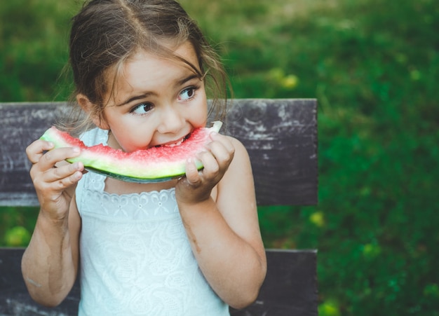 庭でスイカを食べる子供。子供たちは屋外で果物を食べる。子供のための健康的なスナック。スイカのスライスを噛んで庭で遊ぶ少女。