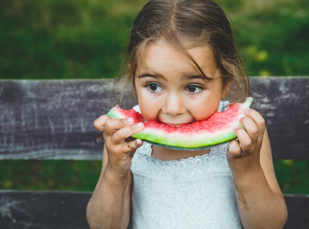 Child eating watermelon in the garden. Kids eat fruit outdoors. Healthy snack for children. Little girl playing in the garden biting a slice of water melon.