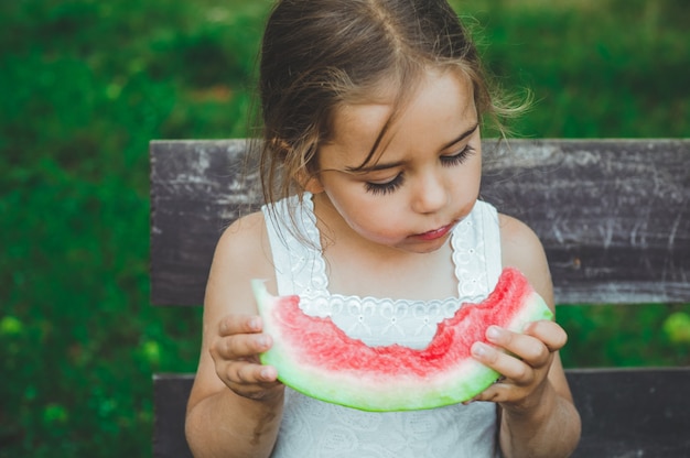Ребенок ест арбуз в саду. дети едят фрукты на открытом воздухе. здоровая закуска для детей. маленькая девочка играет в саду, кусая кусок арбуза.