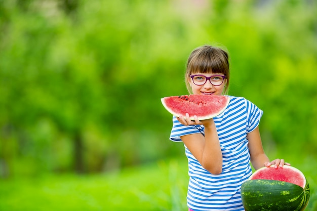 スイカを食べる子供。子供たちは庭で果物を食べます。スイカのスライスを保持している庭の10代前の女の子。スイカを食べる幸せな女の子の子供。ガスと歯のブレースを持つ女児。