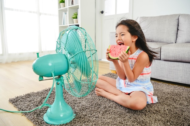 Child eating watermelon with two hands. sitting in front of the electric fan. taking a big bite delightfully