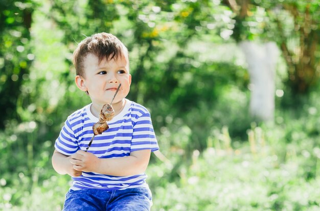 Photo a child eats a kebab on a skewer, in a park or forest nature green background of trees and grass