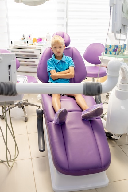 Пятилетний мальчик в синей футболке сидит в сиреневом кресле в стоматологическом кабинете.
