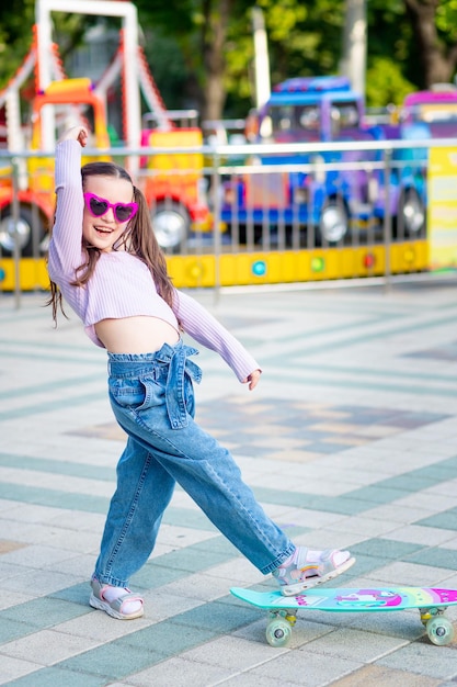 Una bambina in un parco divertimenti cavalca uno skateboard in estate e sorride con felicità vicino alle giostre in occhiali da sole il concetto di vacanze estive e vacanze scolastiche