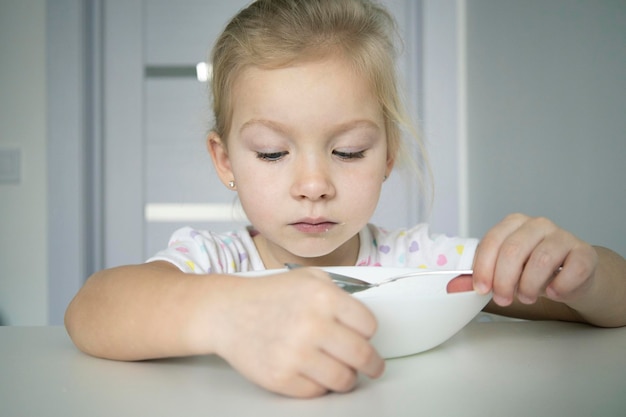 ブロンドの子供の女の子は白いテーブルで白い皿からスプーンで食べる