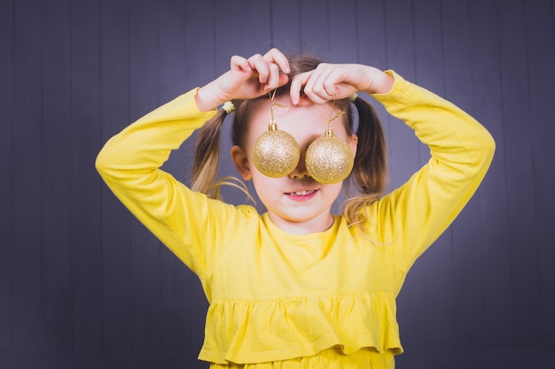 Child girl in bright yellow clothes are playing and having fun with lyellow shiny christmas balls on gray  wooden background