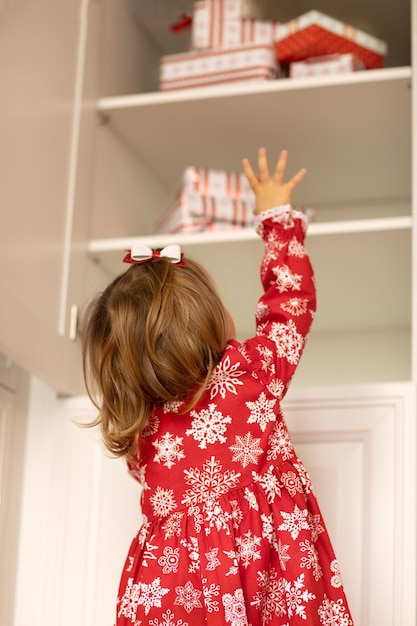 Child girl in christmas dress with snowflakes find gift presents on white cupboard