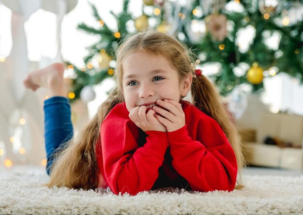 Child girl in Christmas time lying on fluffy carpet and looking at camera in room with decorated tree and gifts. Pretty kid at home in New Year holidays