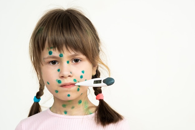 Ребенок, покрытый зеленой сыпью на лице, больной ветряной оспой, корью или вирусом краснухи, держит во рту медицинский термометр