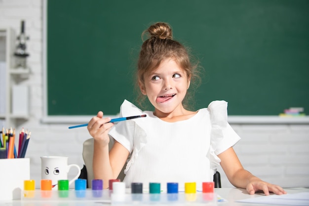 Child girl draws in classroom sitting at a table having fun on school blackboard background little f