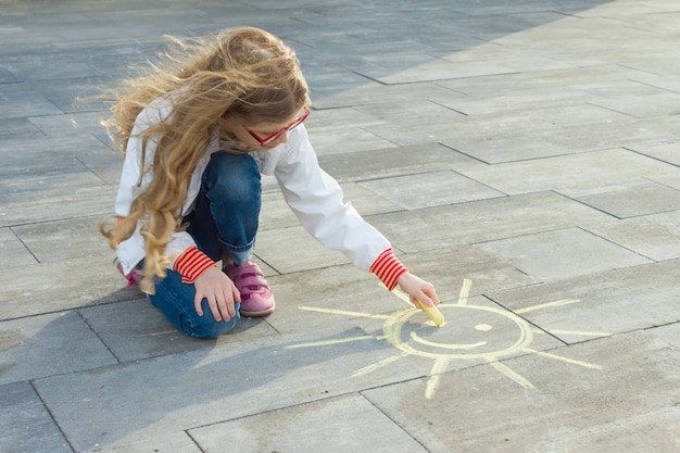 子供の女の子はアスファルトの上に色のクレヨンで太陽のシンボルを描画します。