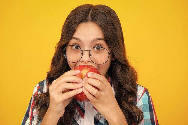 孤立した黄色のスタジオの背景の上にリンゴを食べる子供の女の子果物とtennagerの顔をクローズアップ幸せな面白い笑顔の10代の子供の女の子の肖像画