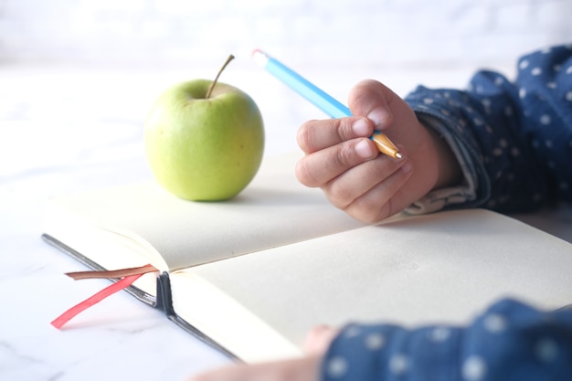 Child girl hand writing on notepad with green fresh apple on table