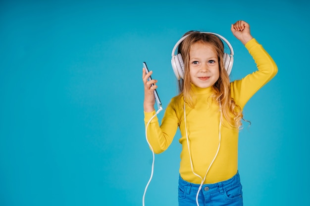 Child girl listen to music on big white headphones and dancing isolated on blue background