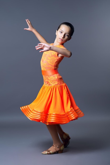 회색 배경에 춤 동작으로 포즈를 취하는 주황색 스포츠 드레스를 입은 어린 소녀
