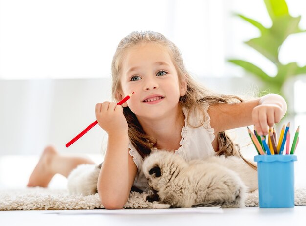 Ragazza del bambino che dipinge con gattini ragdoll sul pavimento e pensa. piccola persona di sesso femminile che disegna con matite colorate e gattini vicino a lei a casa