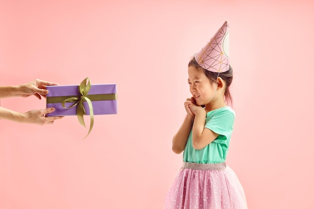 Child Girl in Party Hat Having Feeling of Anticipation Folded her Hands Front Face Looking Gift Box