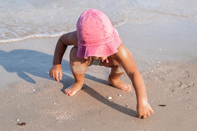 Child girl in a pink panama hat draws on the sand on the beach.