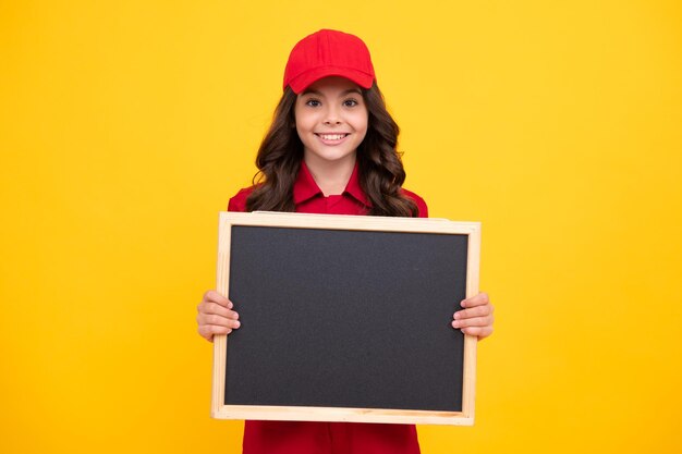 Child girl in red uniform and cap teenage girl hold blackboard isolated on yellow background copy space on empty board mock up happy face positive and smiling emotions of teenager girl