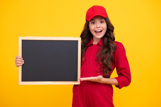 Child girl in red uniform and cap teenage girl hold blackboard isolated on yellow background copy