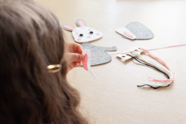 Child girl sews a toy made of felt sitting at the table.