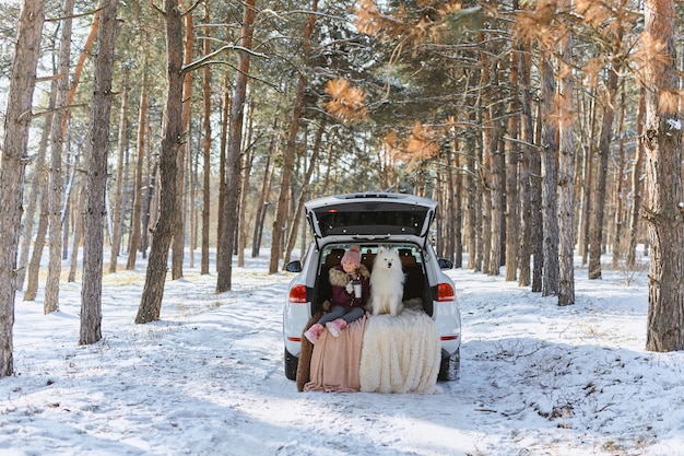 Child girl sitting in the trunk of car with her pet, a white dog Samoyed, in winter in snowy pine forest, a girl drinking tea from a thermos