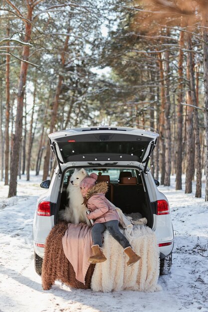 Child girl sitting in the trunk of car with her pet, a white dog Samoyed