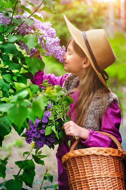 ライラックガーデンガールの麦わら帽子をかぶった子供の女の子は、春のガーデニングで花の香りを吸い込みます