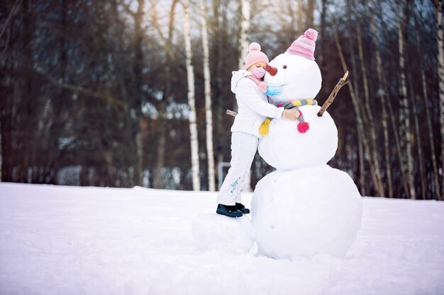 A child girl wearing a mask herself and on a snowman playing with a snowman on a winter walk in nature