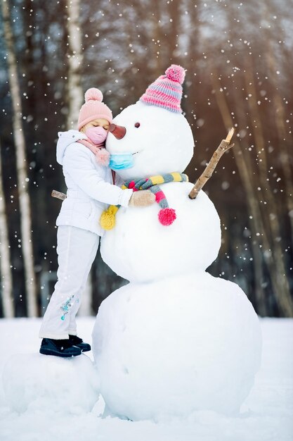 A child girl wearing a mask herself and on a snowman playing with a snowman on a winter walk in nature