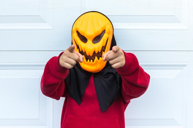 Foto una bambina che indossa una spaventosa maschera di zucca di halloween che punta due dita verso la telecamera sopra una porta di garage bianca concetto di celebrazione costume carnevale terrore paura e autunno