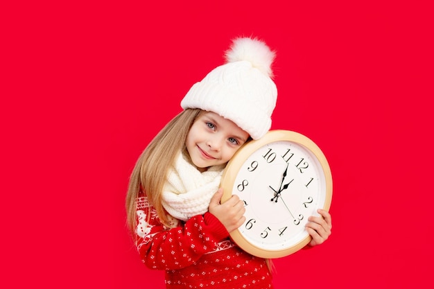 a child girl in a winter hat and sweater with a large clock on a red monochrome isolated background rejoices and smiles the concept of new year and Christmas space for text