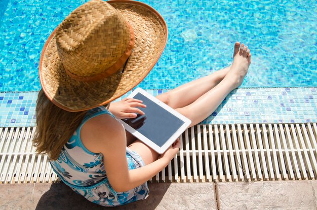 Child girl with big straw hat sitting by the pool using tablet