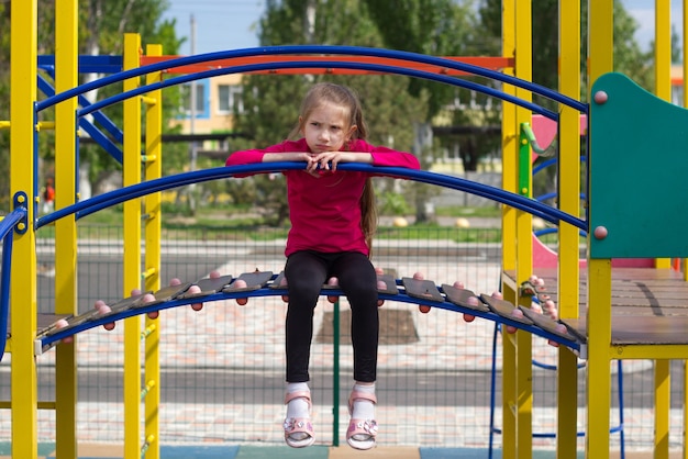 Child girl with blond hair in pink tshirt is sad on the playground sits on slide her legs dangling