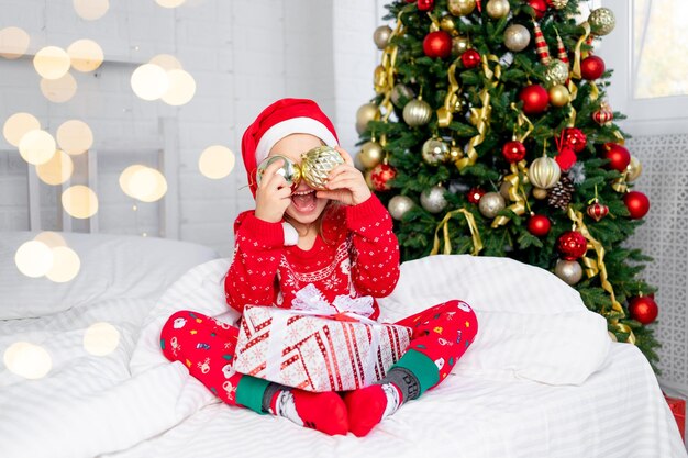 A child girl with Christmas balls on her ears at the Christmas tree in a red sweater and Santa Claus hat on New Year's Eve in a white bed smiling, fooling around and laughing with a gift