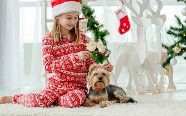 Child girl with dog at Christmas time at home with holiday decoration. Kid wearing red costume and pet doggy in room with New Year decoration