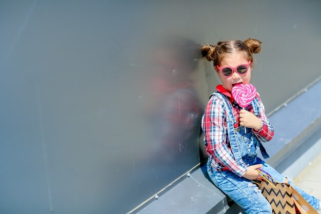 A child a girl with a Lollipop in her hands sits on the street in a denim jumpsuit and sunglasses