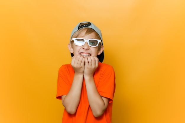 A child in glasses for watching movies bites his nails out of fear