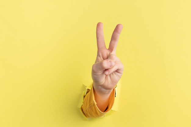 Child hand counting and showing two finger up through hole in\
yellow paper with torn edges peace gesture or victory v sign
