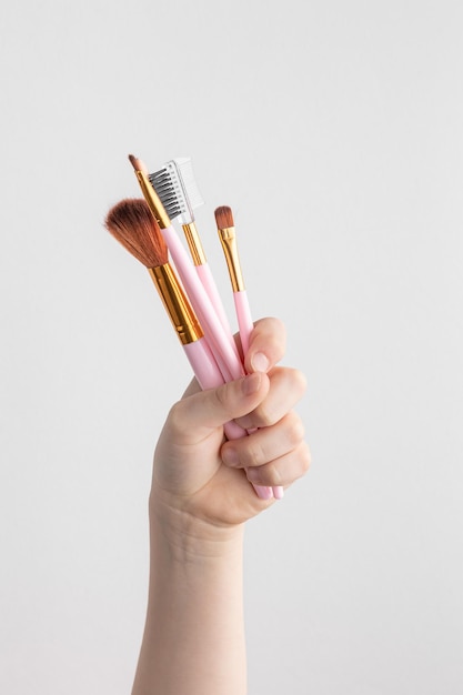 Child hand holding many pink makeup brushes above a white and gray paper background copy space