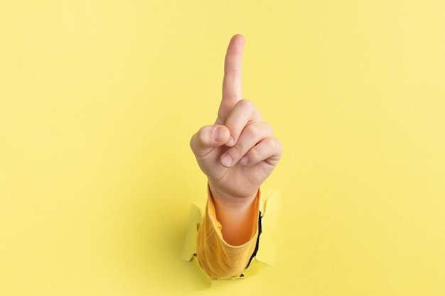 Child hand shows the forefinger up through hole in yellow paper with torn edges concept press the button first