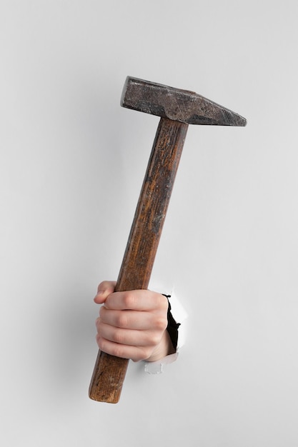 Photo child hand through hole in white paper with torn edges holding a hammer copy space