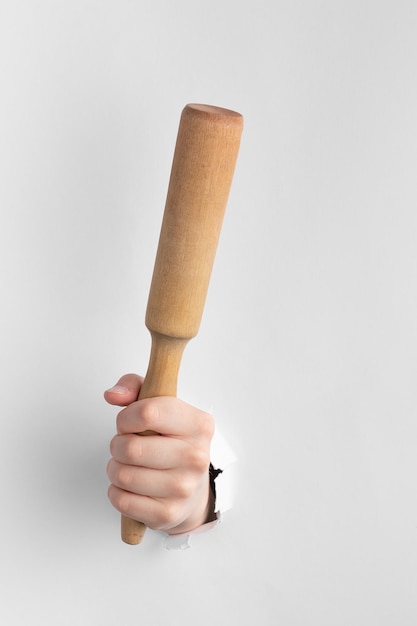 Child hand through hole in white paper with torn edges holding a wooden rolling pin kitchen tool for cooking above a paper background