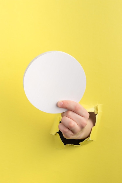 Child hand through hole in yellow paper with torn edges holding a white round wooden block copy space