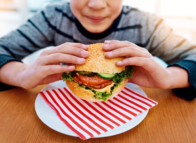 Child hands and eating burger on table for delicious lunch meal or food with healthy vegetables at home Hand of hungry little boy holding beef hamburger for fresh dinner nutrition or vitamins