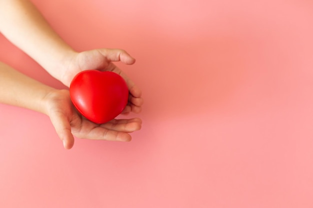 Child hands holding red heart on pink background