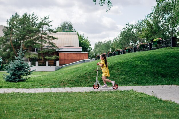 A child has a good time riding a scooter on a path in the city between green grass and trees