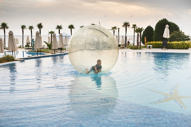 Ребенок развлекается внутри большого пластикового шара на воде бассейна на летнем курорте. Маленький мальчик внутри большой надувной прозрачный мяч работает и развлекается.