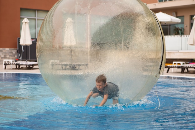 Child have fun inside big plastic balloon on the water of\
swimming pool on the summer resort. little boy inside big\
inflatable transparent ball running and having fun.