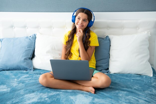 Child in headphones relax on bed at home listen to music using laptop Child in earphones browse internet on computer